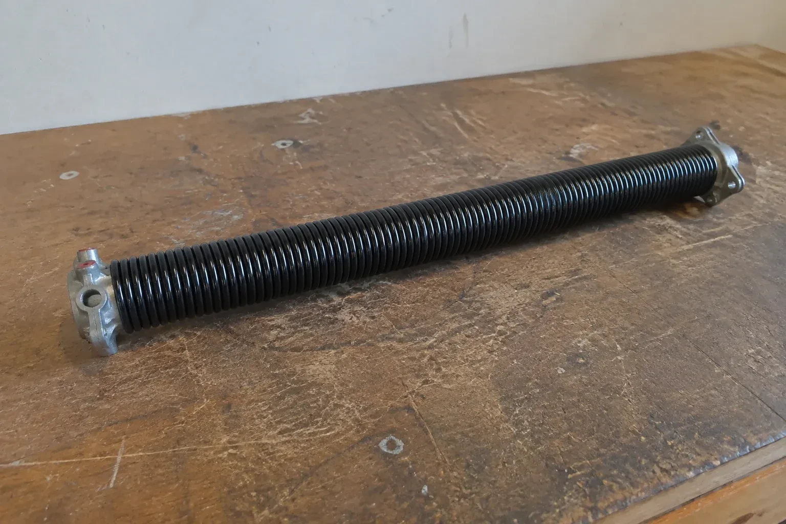 Detached black coil spring with metal end fittings on a workbench. — Torsion Spring Replacement, Hamilton