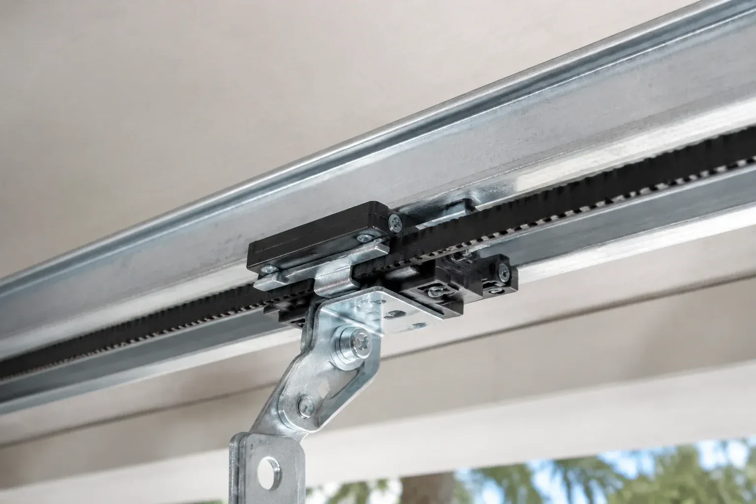 Black plastic trolley and bent galvanized bracket on a steel opener rail with a toothed black belt, against a ceiling. — Belt-Drive Opener Repair, Hamilton